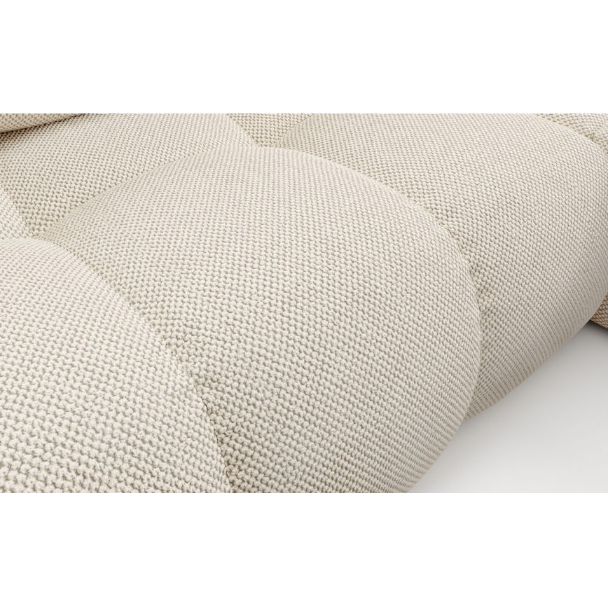 Čalúnený nábytok s jemnou textúrou látky je umiestnený na svetlom pozadí. Látka je svetlo béžová s viditeľnou textúrou pripomínajúcou pletenie. Jednotlivé časti nábytku sú prešívané do mäkkých vankúšovitých tvarov.