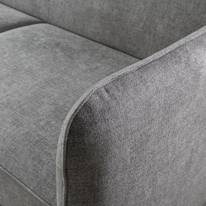 Sivá pohovka s mäkkým čalúnením, zaoblenými lakťovými opierkami a jemnou štruktúrou látky. Je zhotovená z textílie s krátkym vlasom.