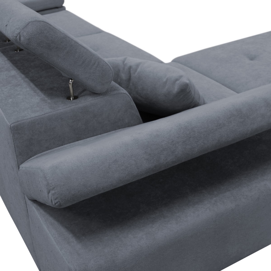 Rohová sedačka sivomodrej farby je zachytená zblízka. Sedačka má podhlavníky a valcovité opierky. Je fotografovaná na bielom pozadí.