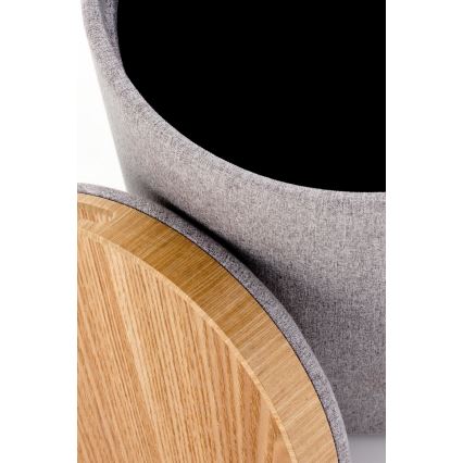 Šedá látková úložná taburetka s dreveným vekom je čiastočne otvorená, odhaľujúc vnútorný priestor. Veko je z ľahkého dreva s okrúhlym tvarom. Taburetka je umiestnená na svetlom pozadí.