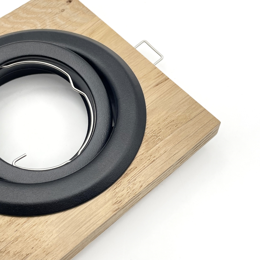 Čierny okrúhly rámček s vnútorným kovovým držiakom je pripevnený k drevenej doske. Rámček je umiestnený v strede dosky, ktorá má svetlohnedú farbu. Na pravom hornom rohu drevenej dosky je malý kovový háčik.