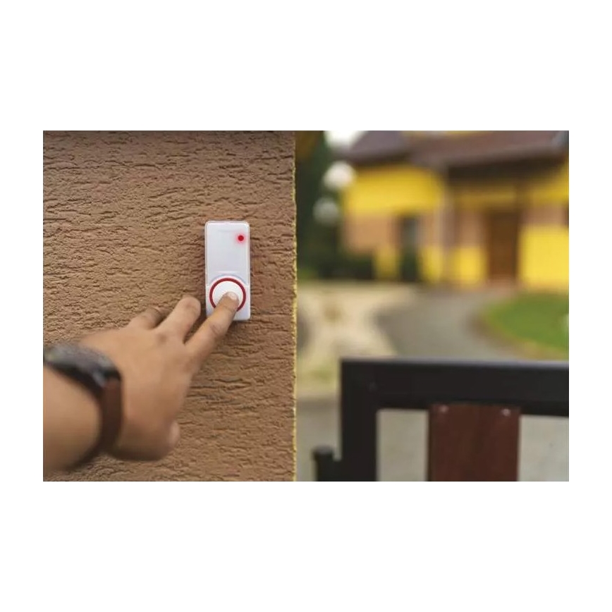 Ruka stláča tlačidlo bezdrôtového zvončeka pripevneného na hnedú stenu. V pozadí je viditeľná časť záhrady a žltý dom.