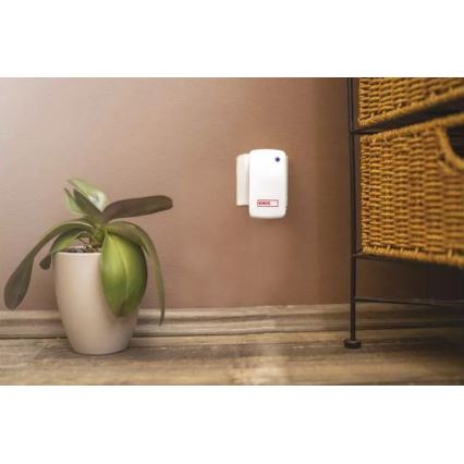 Biely odpudzovač hmyzu je pripevnený na stene. Nachádza sa vedľa kvetináča s rastlinou a drevenej komody. Na odpudzovači je malé červené logo „4MOS“.