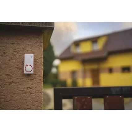 Zvonček je pripevnený na hnedom múre. Nachádza sa vonku pred žltým domom s bránou.