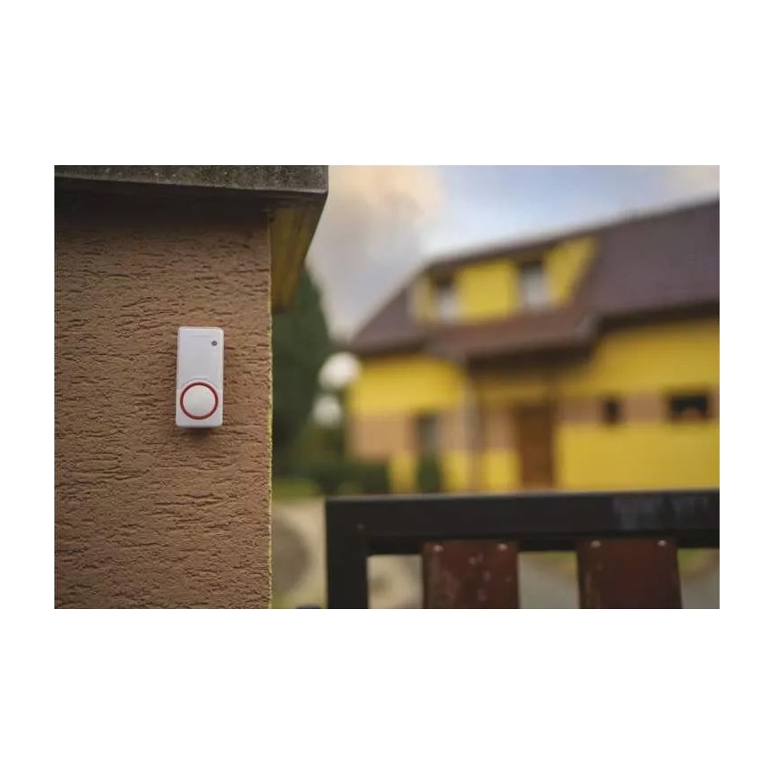 Zvonček je pripevnený na hnedom múre. Nachádza sa vonku pred žltým domom s bránou.