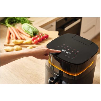 Čierna fritéza Sencor s dotykovou obrazovkou, na ktorej je nastavená teplota 180°C a čas 20 minút, je ovládaná rukou. Na stole pred ňou je drevená doska s nakrájanou zeleninou.