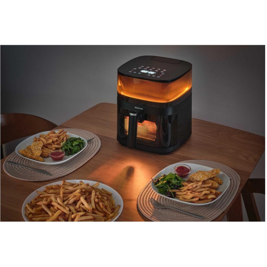 Čierna fritéza s osvetleným vnútrom pripravuje jedlo. Na drevenom stole sú dve taniere s hranolkami a kuracími prúžkami.