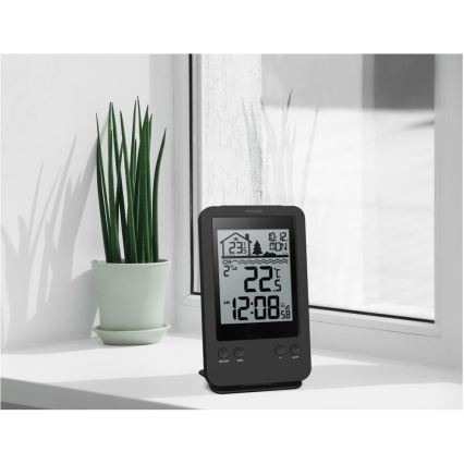 Digitálna meteostanica zobrazuje čas 12:08, teplotu 22,5 °C a vlhkosť 58 %. Stojí na parapete pred oknom vedľa kvetináča s rastlinou.