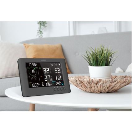 Digitálna meteostanica zobrazuje čas, teplotu, vlhkosť a atmosférický tlak. Stojí na bielom konferenčnom stolíku v obývacej izbe s pohovkou a kvetináčom.