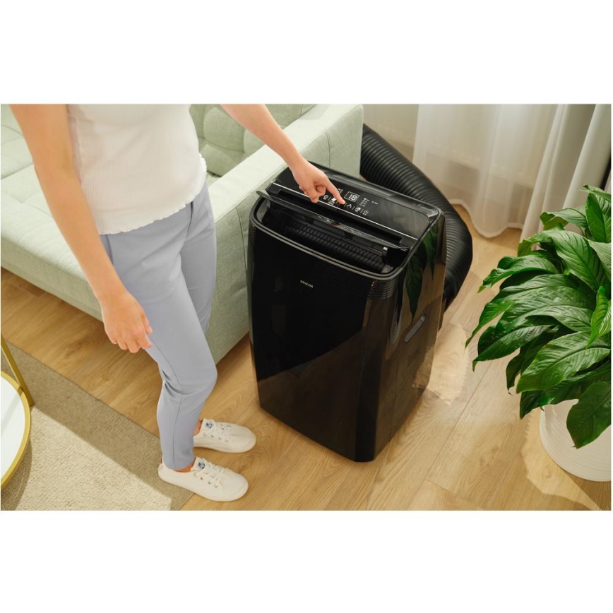 Mobilná klimatizácia s čiernym lesklým povrchom stojí na drevenej podlahe obývačky. Žena nastavuje jej ovládací panel. V okolí je pohovka a izbová rastlina.