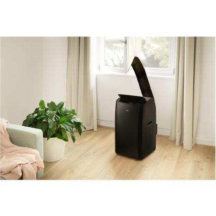 Mobilná klimatizácia stojí na svetlej drevenej podlahe v obývačke. Zariadenie je čierne, s vývodom odvádzajúci teplý vzduch cez okno. V miestnosti je pohovka, kvetináč s rastlinou a závesy.