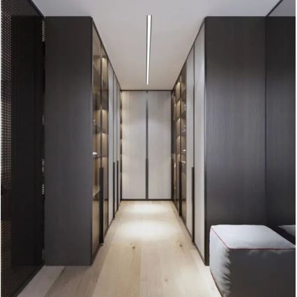 Šatník s tmavými skriňami a presklenými policami sa rozprestiera po oboch stranách dlhej chodby s svetlou drevenou podlahou a stropným osvetlením. Na konci chodby je svetlý panel. Pri stene je umiestnený mäkký sedací puf.