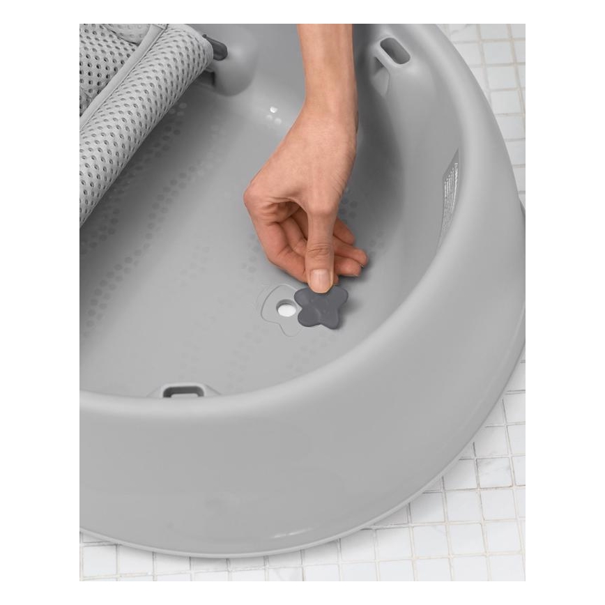 Ruka vkladá štuple do odtoku detskej vane šedej farby. Vaňa je umiestnená na dlaždicovej podlahe.