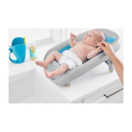 Dojča leží v šikmej vaničke na umývadle. Dospelá osoba umýva dojča. Vedľa vaničky je modrá naberačka a fľaštička s tekutinou.