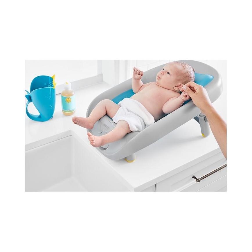 Dojča leží v šikmej vaničke na umývadle. Dospelá osoba umýva dojča. Vedľa vaničky je modrá naberačka a fľaštička s tekutinou.