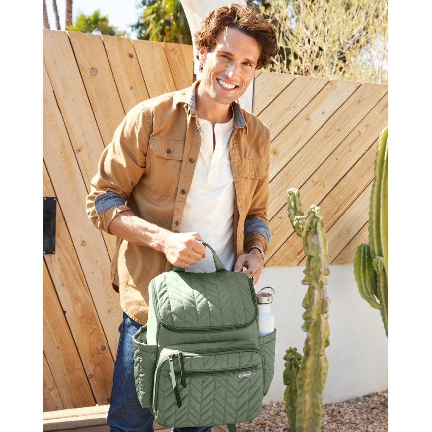 Muž balí olivovozelenú prebaľovaciu tašku s prešívaným vzorom. Do bočného vrecka vkladá fľašu. Stojí pred dreveným plotom s kaktusmi.
