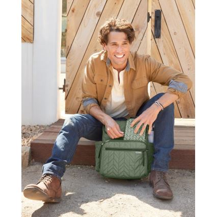 Muž v hnedom kabáte a džínsoch sedí pri drevených dverách a ukladá predmety do olivovo zelenej prešívanej tašky. Taška má zips a vrecko s grafickým vzorom.