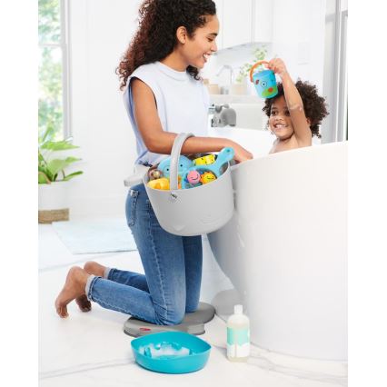 Žena na kolenách drží vedro s kúpeľovými hračkami pri vani, v ktorej sa hrá malé dieťa. Vedro je sivé, hračky sú modré a žlté. Na zemi je modrá miska a fľaša šampónu.