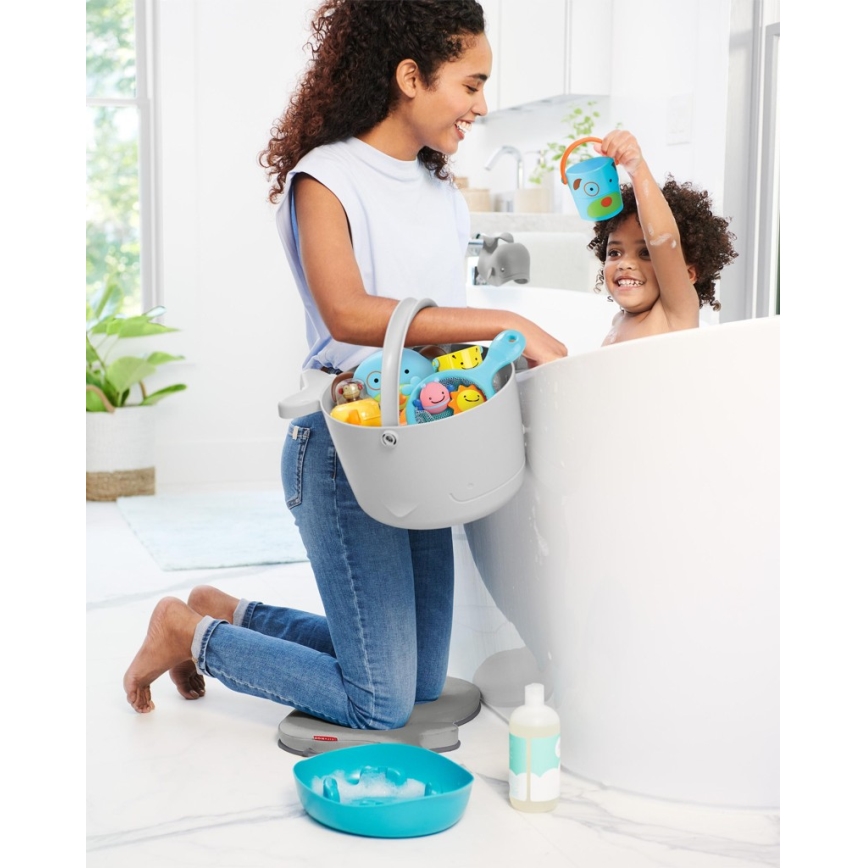 Žena na kolenách drží vedro s kúpeľovými hračkami pri vani, v ktorej sa hrá malé dieťa. Vedro je sivé, hračky sú modré a žlté. Na zemi je modrá miska a fľaša šampónu.