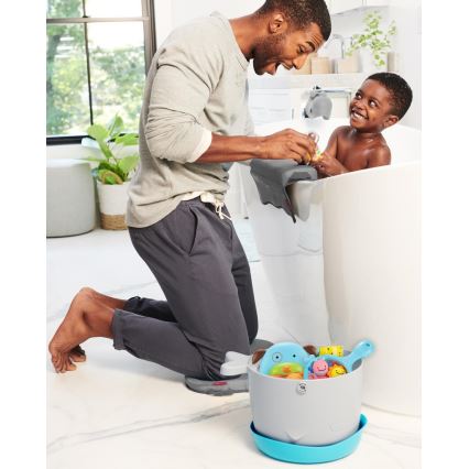 Otec kúpe malého syna v vani. Otec kľačí pri vani a drží opierku na kolená. V blízkosti je vedro s detskými kúpacími hračkami.