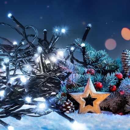 Elektrický svetelný veniec s bielymi LED diódami leží na snehu popri vianočnej jedli zdobenej šiškami a bobuľami. Vedľa je drevená hviezdička.