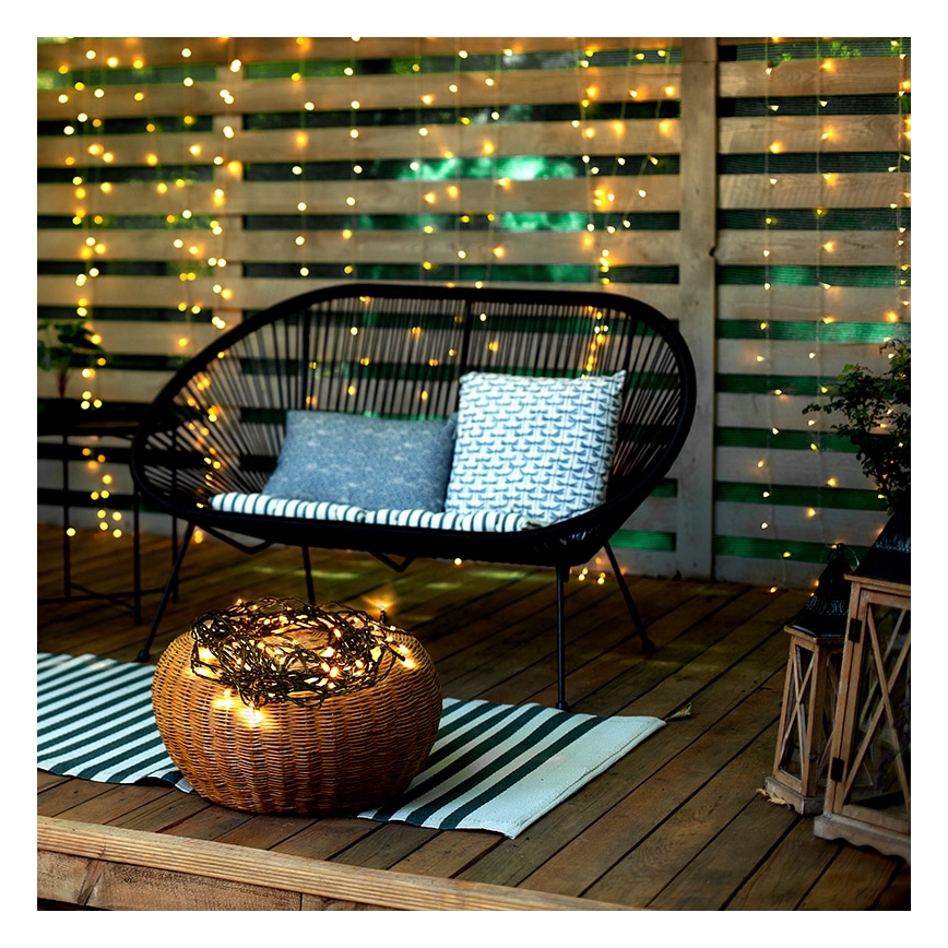 Pletaná nádoba s rozžiarenými svetielkami stojí na terase pred čiernou záhradnou lavicou. Lavica je umiestnená pred drevenou stenou ozdobenou svetielkami.