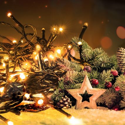 Vianočné svetielka svietia v tmavšom prostredí. Sú umiestnené na umelom snehu medzi vetvičkami ihličnatého stromčeka a drevenou hviezdou.