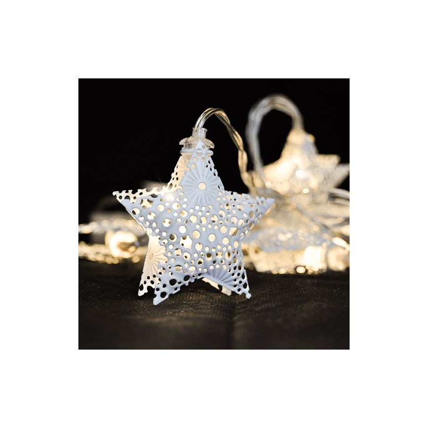 Biele kovové hviezdy s prelamovaným vzorom svietia. Sú pripevnené na svetelnom reťazi. Ležia na tmavej ploche.