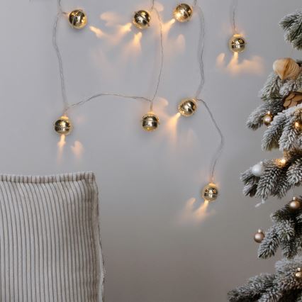 Svetlený veniec s malými zlatými zvončekmi visí na stene. Svetlá svietia. V blízkosti je čiastočne viditeľný vankúš a zasnežený vianočný stromček.