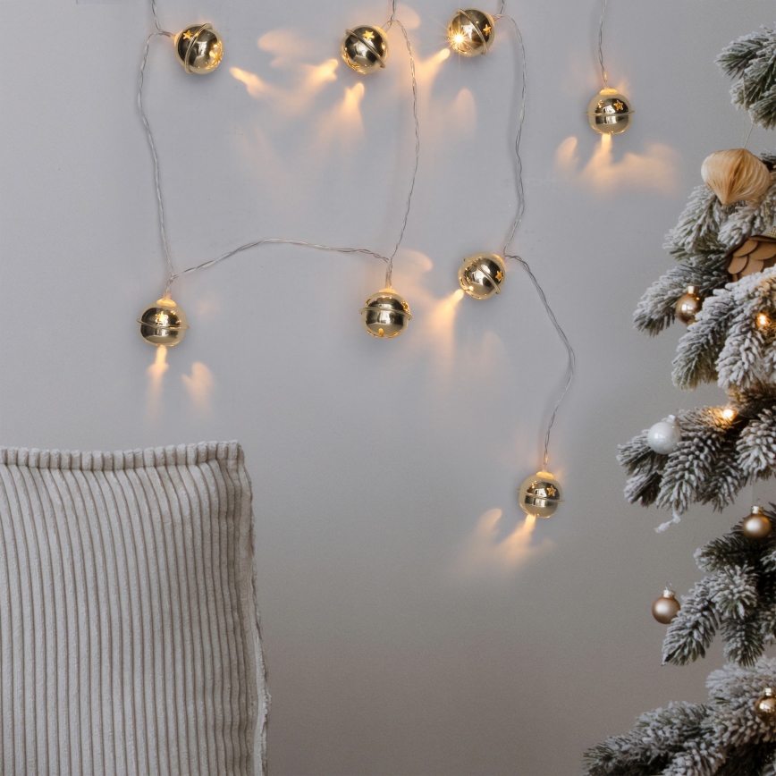 Svetlený veniec s malými zlatými zvončekmi visí na stene. Svetlá svietia. V blízkosti je čiastočne viditeľný vankúš a zasnežený vianočný stromček.