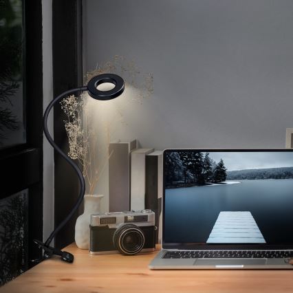 Stolná lampa s kruhovou hlavou svieti na drevenom stole. Je pripevnená sponou. Na stole je fotoaparát a notebook s obrázkom zasneženej krajiny.