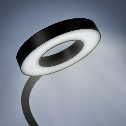 Kruhová stolová lampa svieti. Je umiestnená na ohybnom rameni pred svetlosivým pozadím.