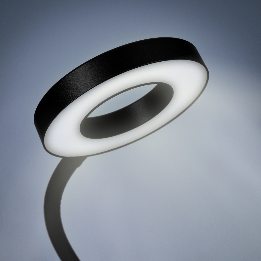Kruhová stolová lampa svieti. Je umiestnená na ohybnom rameni pred svetlosivým pozadím.