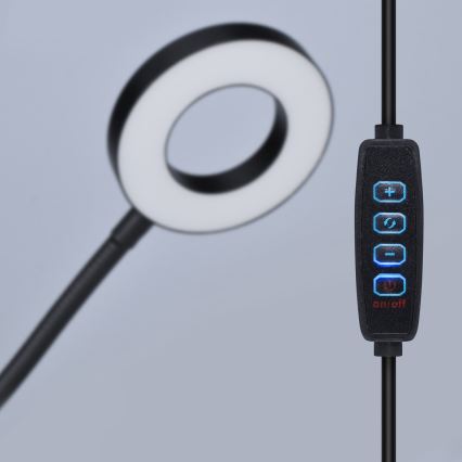 Stolná lampa s kruhovým svetelným zdrojom je umiestnená na stole. Na kábli je ovládač s tlačidlami: +, šípka do kruhu, -, a tlačidlom on/off.