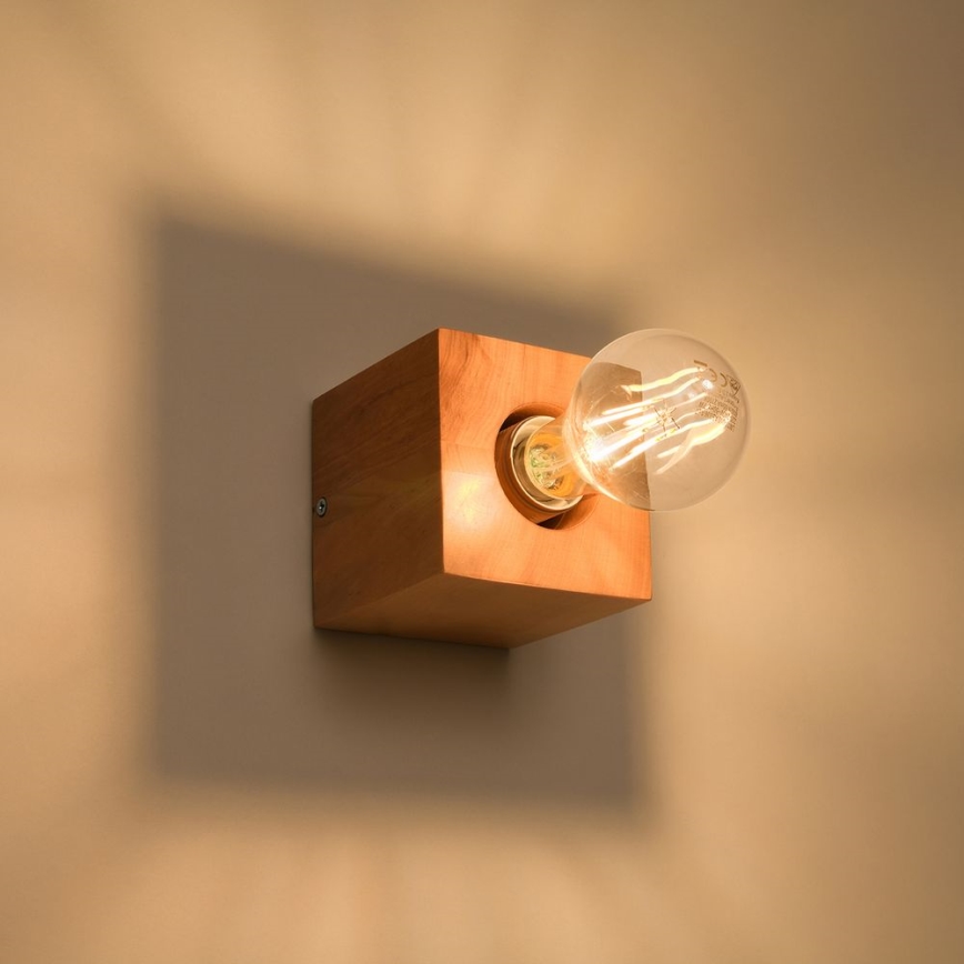 Drevená nástenná lampa svieti. Žiarovka je zasunutá do otvoru v kubickej drevenej konštrukcii. Lampa je pripevnená na stene.