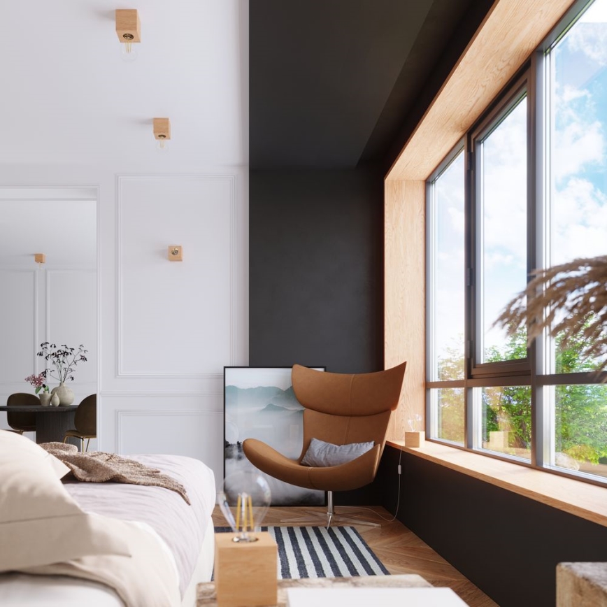 Hnedé kreslo stojí v rohu miestnosti s tmavou stenou a veľkým oknom. Miestnosť je svetlá, s bielymi stenami a drevenými detailmi. Vedľa kresla je posteľ a stolík.