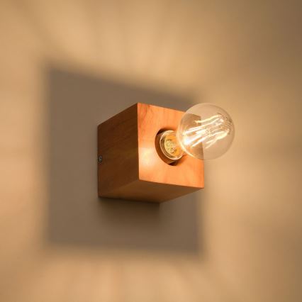 Drevená nástenná lampa svieti. Žiarovka je zasunutá do otvoru v kubickej drevenej konštrukcii. Lampa je pripevnená na stene.