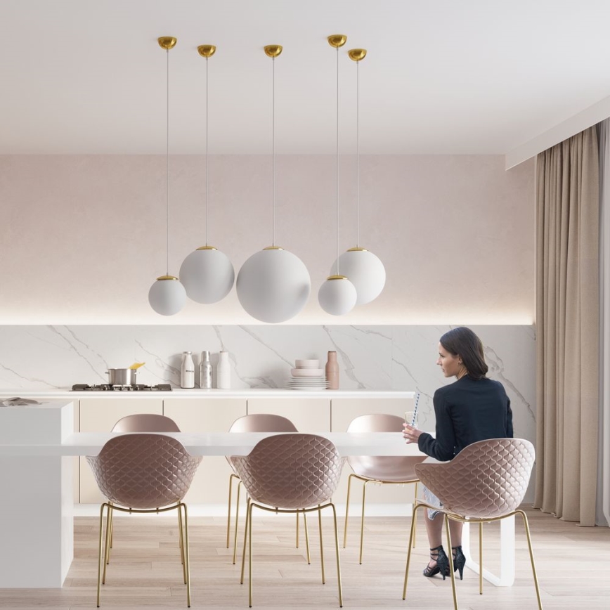 Žena sedí pri bielom jedálenskom stole s ružovými stoličkami v moderne zariadenej kuchyni. Nad stolom visí niekoľko bielych guľových svietidiel so zlatými detailmi. Kuchyňa má mramorovú pracovnú dosku a béžové steny.