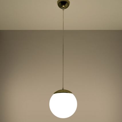 Guľová lampa visí zo stropu na tenkom kábli. Lampa má bielu guľovú časť a zlatý kovový vrchný a spodný okraj. Strop a stena sú svetlohnedé.