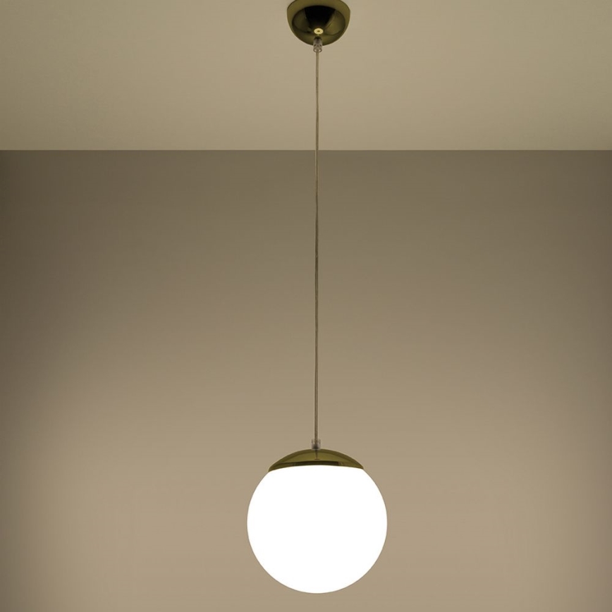 Guľová lampa visí zo stropu na tenkom kábli. Lampa má bielu guľovú časť a zlatý kovový vrchný a spodný okraj. Strop a stena sú svetlohnedé.
