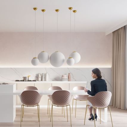 Žena sedí pri jedálenskom stole a číta dokumenty. Stôl je obklopený ružovými stoličkami s kovovými nohami. Nad stolom visí päť bielych guľových svietidiel s mosadznými detailmi.