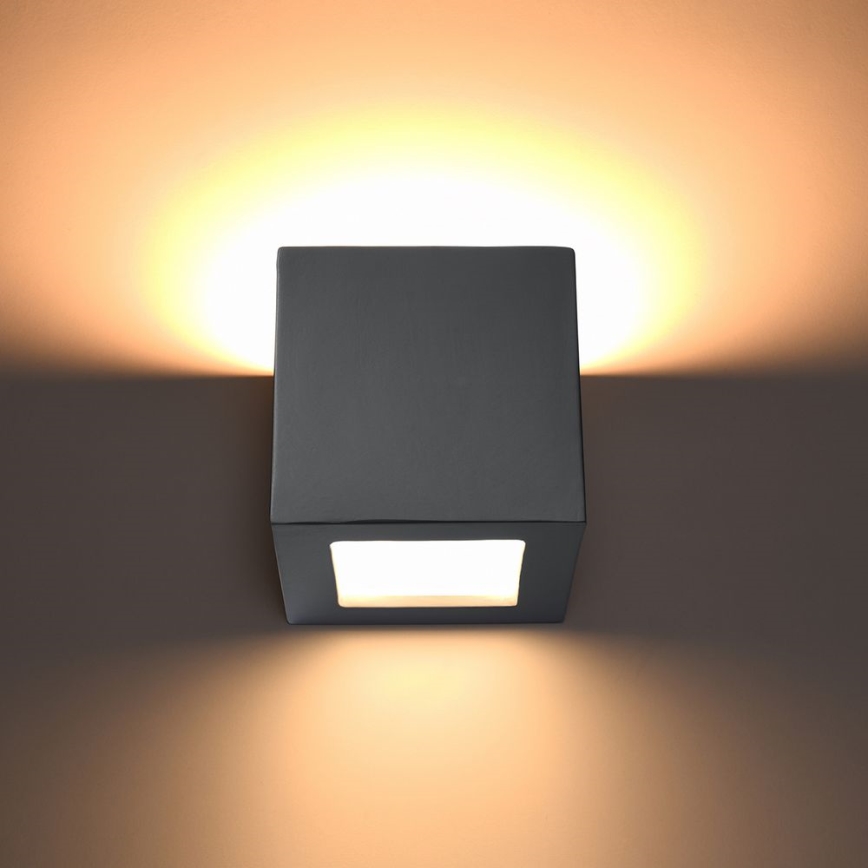 Kocková nástenná lampa svieti na stene. Svetlo vyžaruje z otvoru v strede lampy, osvetľujúc okolité steny.
