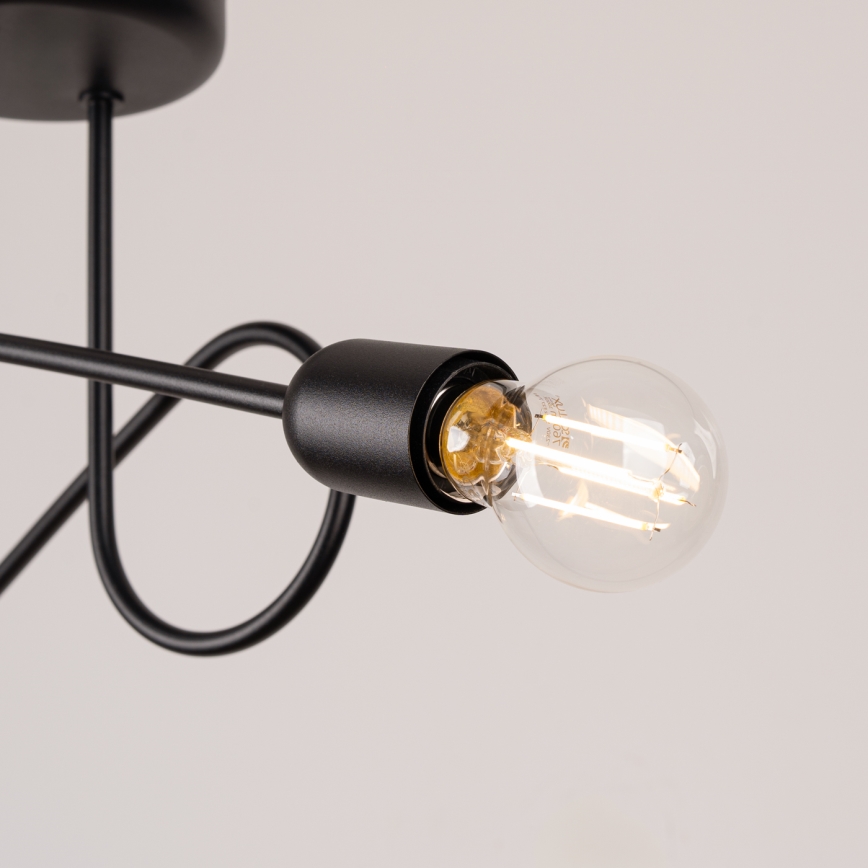 Čierna stropná lampa s jednou žiarovkou, tvarovaná do oblúka, je pripevnená k stropu. Žiarovka je zapustená do čiernej objímky. Svetlo svieti na neutrálne sivé pozadie.