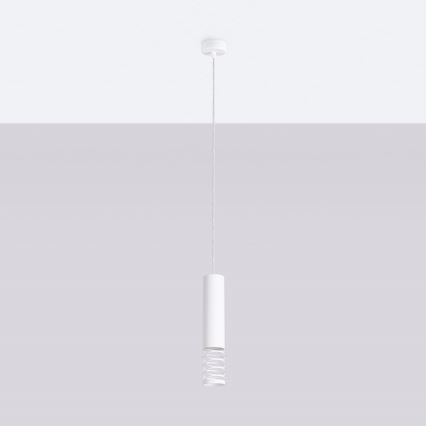 Biela závesná lampa visí zo stropu. Lampa má valcovitý tvar s dekoratívnym pružinovým prvkom. Je umiestnená pred svetlosivou stenou.