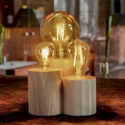Tri žiarovky – jedna veľká a dve menšie – osvetlené, umiestnené na drevených podstavcoch. Podstavce sú usporiadané vedľa seba na tmavom drevenom stole.
