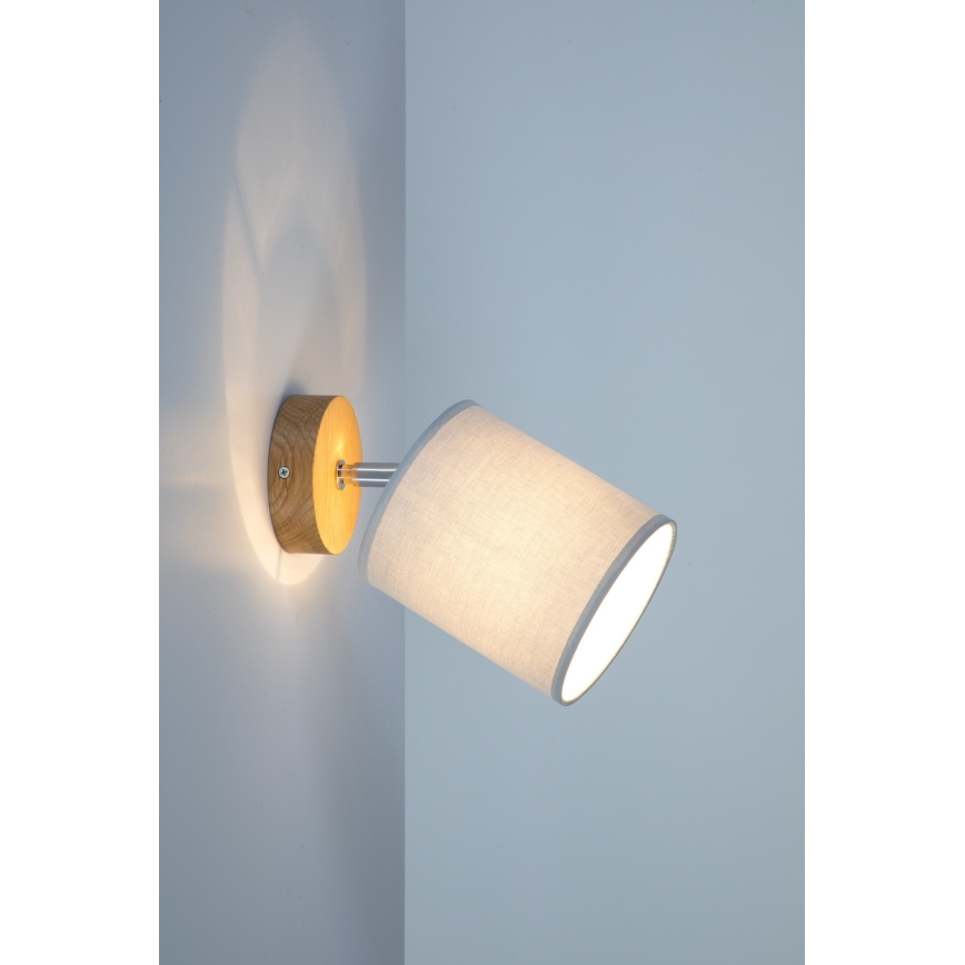 Nástenná lampa so svetlošedým textilným tienidlom a drevenou podstavou svieti na svetlošedkú stenu.
