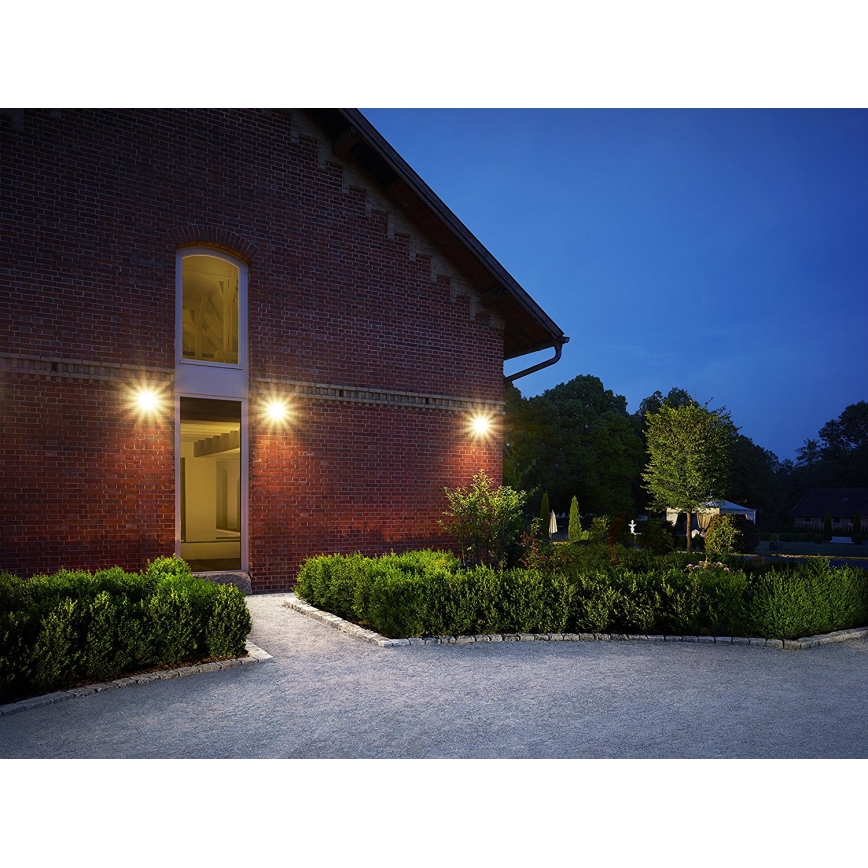 Tri vonkajšie nástenné svetlá osvetľujú tehlovú budovu v noci. Svetlá sú umiestnené symetricky na stene pri vchode. Okolitá záhrada je upravená, s nízkymi živými plotmi.