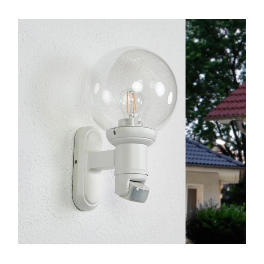 Biela nástenná lampa s guľovou sklenenou clonou a zabudovaným pohybovým senzorom je namontovaná na stene budovy. Lampa obsahuje žiarovku. Na pozadí je viditeľná časť záhrady s domom.
