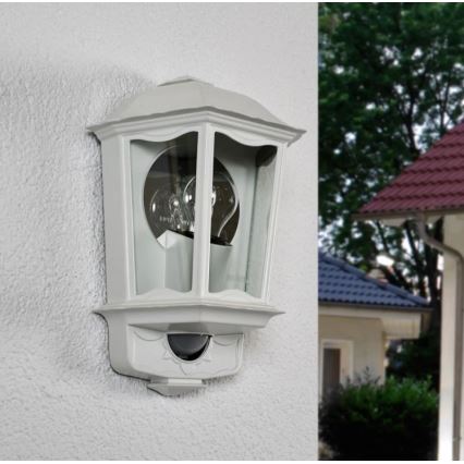 Svetlo s pohybovým senzorom je namontované na vonkajšej stene. Je to sivá, klasicky tvarovaná nástenná lampa. V pozadí je vidieť časť rodinného domu a zeleň.