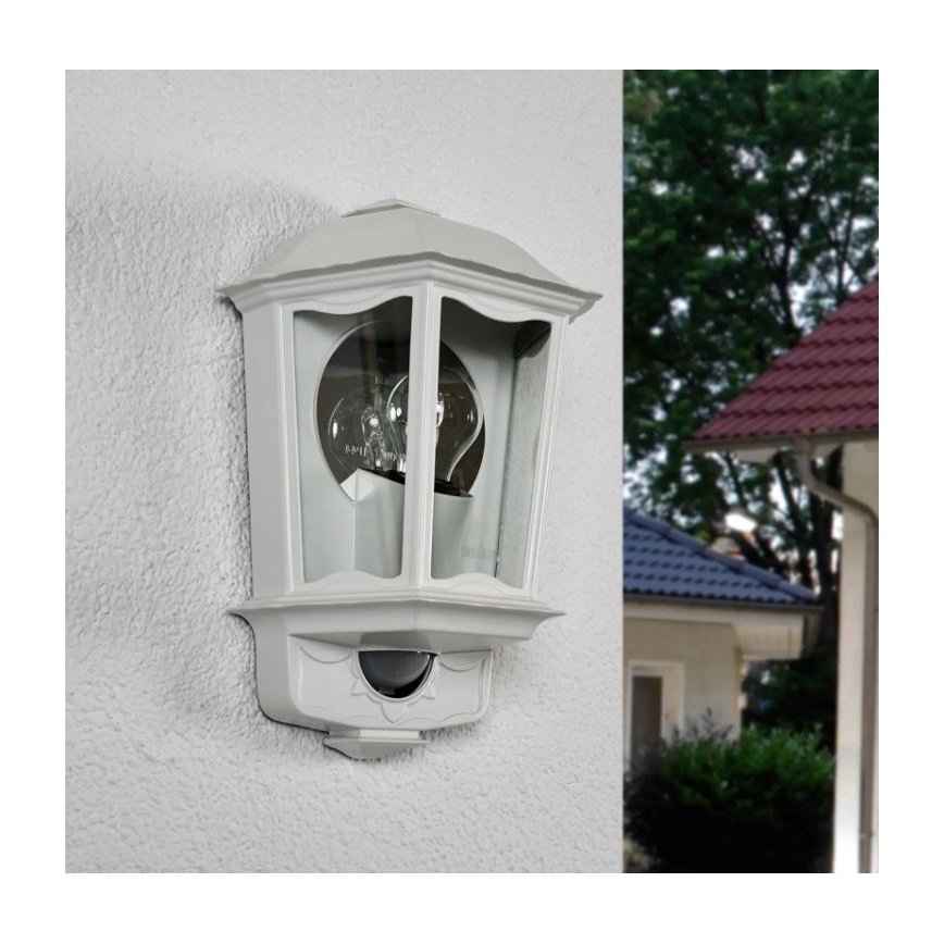 Svetlo s pohybovým senzorom je namontované na vonkajšej stene. Je to sivá, klasicky tvarovaná nástenná lampa. V pozadí je vidieť časť rodinného domu a zeleň.